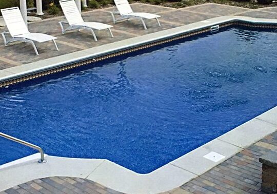 A swimming pool with two white chairs and a brick floor.